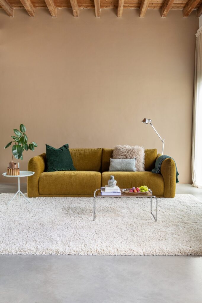 Okergele bank Lova van By SIDDE in een warme woonkamer met houten balken, plant en hoogpolig vloerkleed